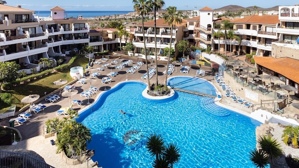 Ein Resort mit einem zentral gelegenen Schwimmbad, umgeben von Liegestühlen und Palmen, mit mehreren Hotelgebäuden im Hintergrund.