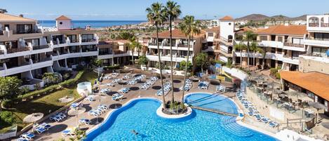 A resort with a central swimming pool surrounded by sun loungers and palm trees, with multiple hotel buildings in the background.