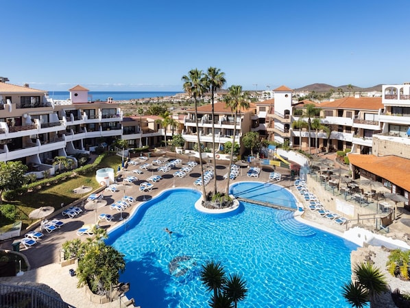 Ein Resort mit einem zentral gelegenen Schwimmbad, umgeben von Liegestühlen und Palmen, mit mehreren Hotelgebäuden im Hintergrund.