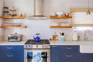 A kitchen with a stove, oven, and a blue kettle. There are shelves with glass jars and a toaster. - Charming Renovated Cottage, Perfect Location, Walk to Everything, Across From Park & Cultural Center (Newberg)