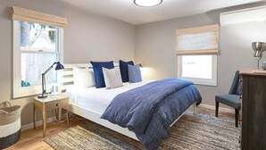 A bedroom with a bed, bedside table, lamp, chair, and a window with blinds. - Charming Renovated Cottage, Perfect Location, Walk to Everything, Across From Park & Cultural Center (Newberg)
