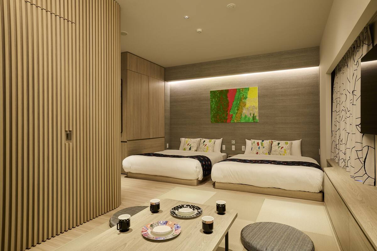 A modern hotel room with two beds, a small table with plates and cups, a colorful abstract painting on the wall, and a TV on the right side.
