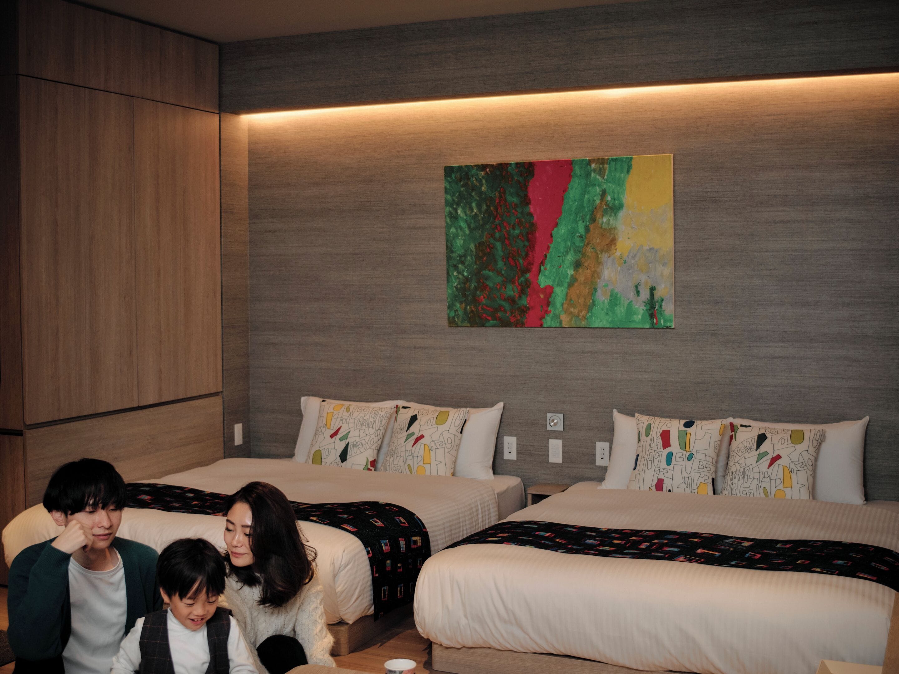 A modern hotel room with two beds, a large abstract painting on the wall, and a wooden wardrobe.