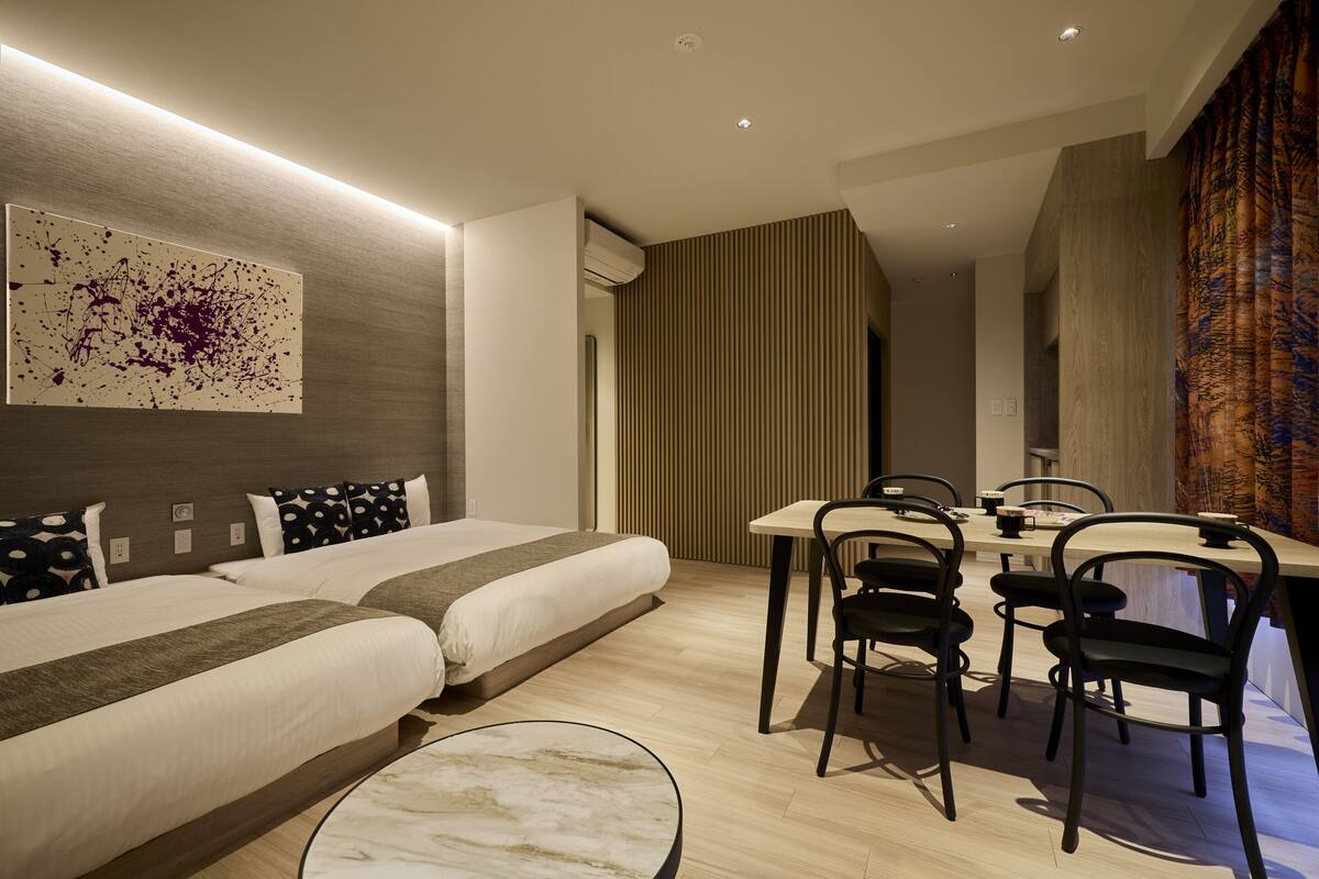A modern hotel room with two beds, a dining area with chairs, and a large abstract painting on the wall.
