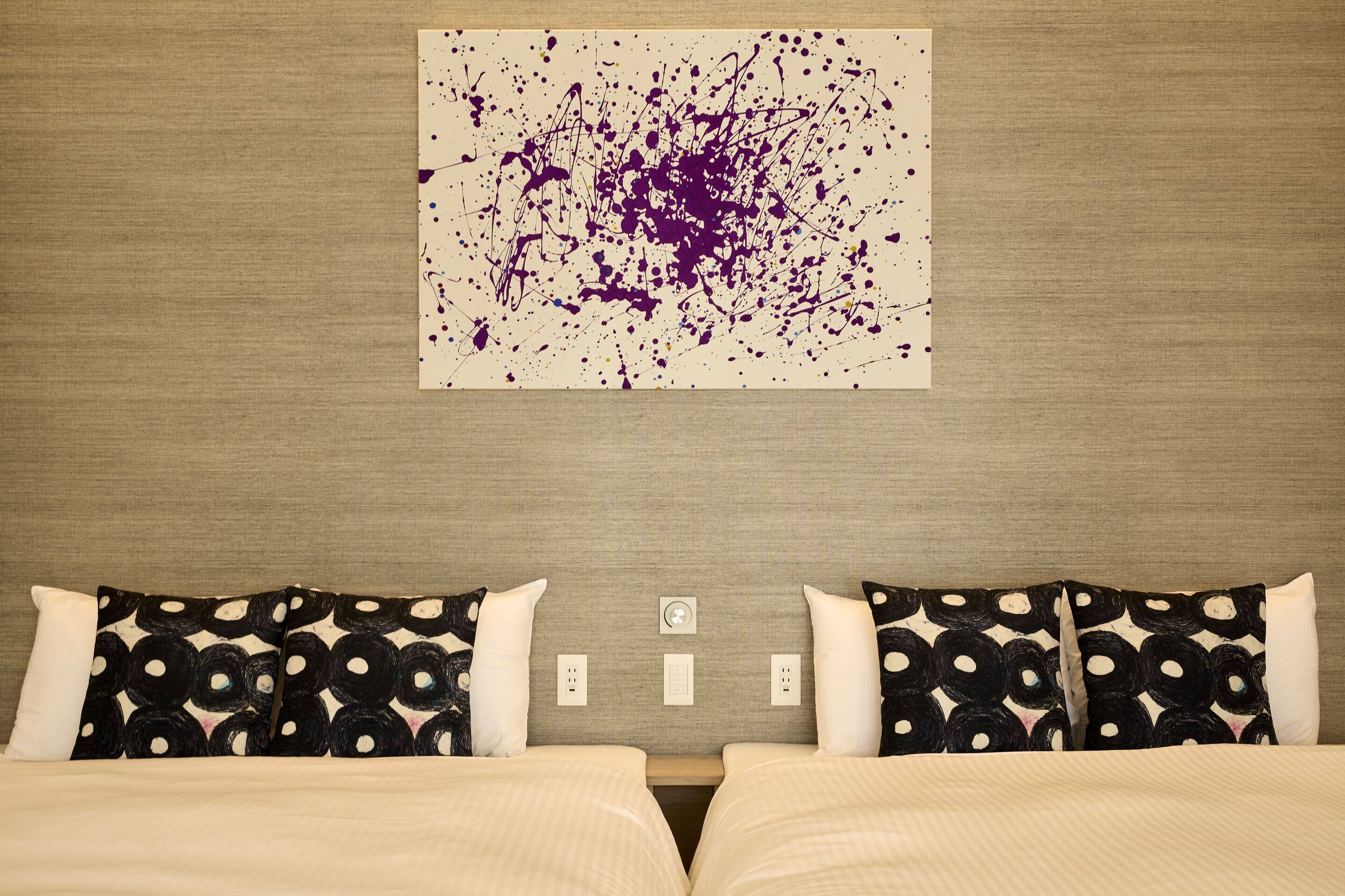 Habitación de hotel con una cama, dos almohadas con estampado blanco y negro, un cuadro abstracto colgado en la pared y un enchufe.
