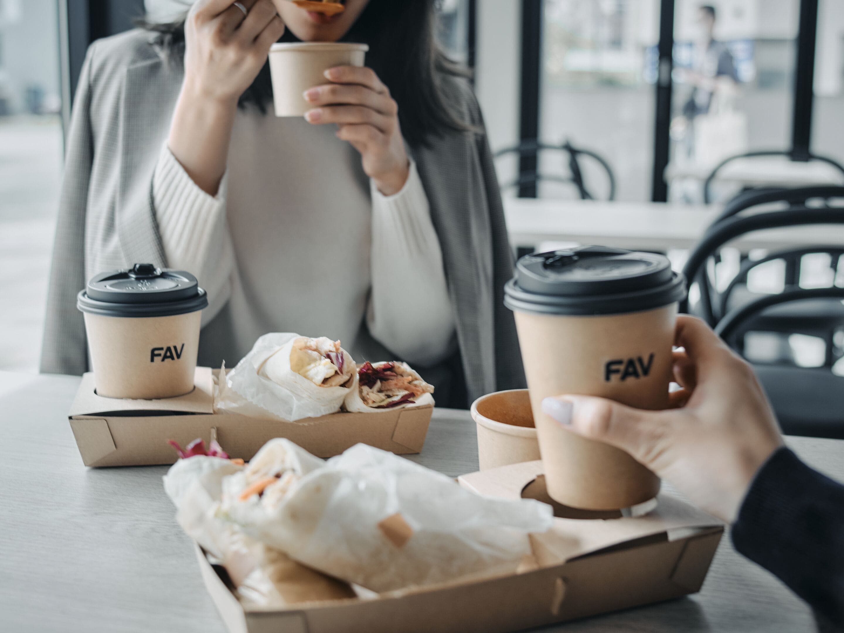 Une personne tient une tasse de café avec un couvercle, un sandwich dans une boîte en papier, et une autre tasse de café est posée sur une table.