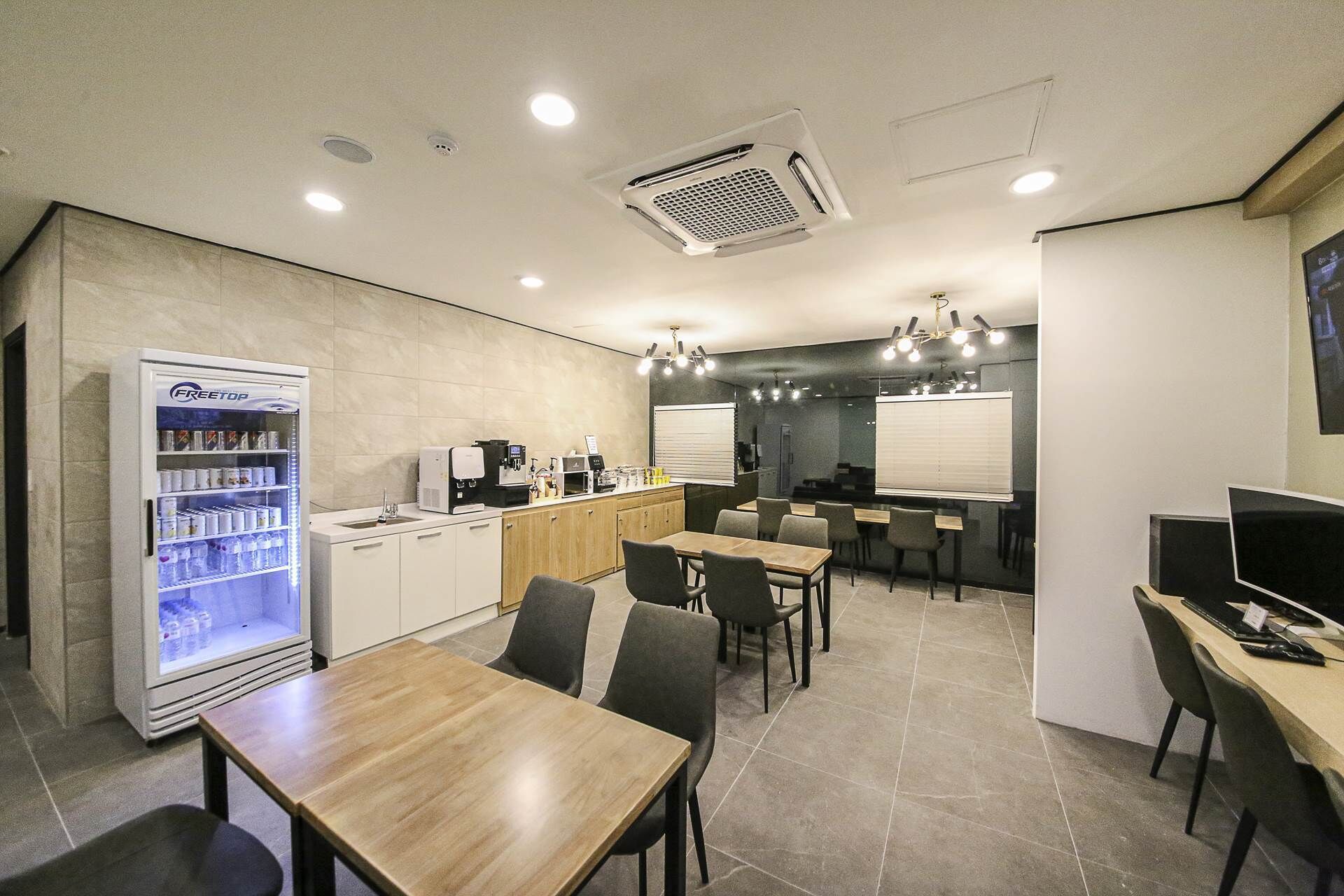 A modern cafe with a freestanding beverage dispenser, a coffee machine, and a seating area with tables and chairs. - Arden Hotel (Cheonan)