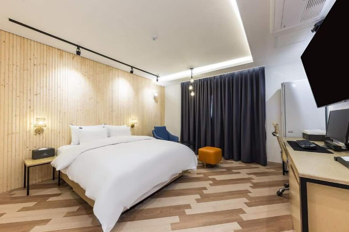 A modern hotel room with a wooden panel wall, a large bed, a desk with a computer, and a chair.