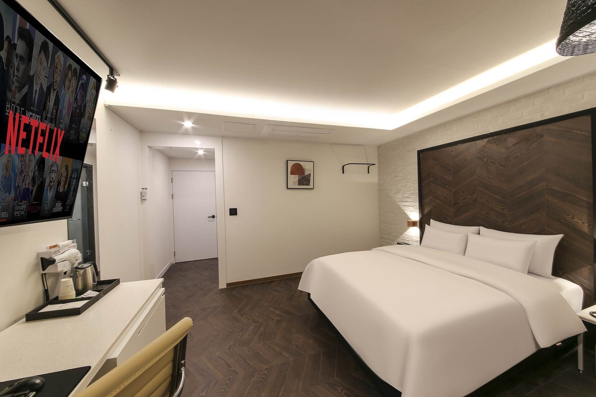 A hotel room with a large bed, a desk with a computer, a chair, a TV mounted on the wall, and a door leading to another room.