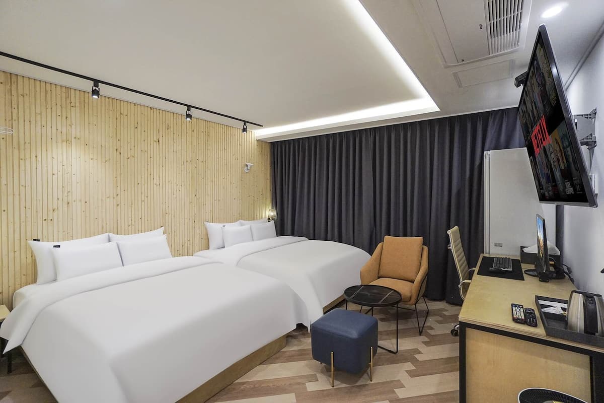 A hotel room with two beds, a desk with a chair, a TV, and a small ottoman.