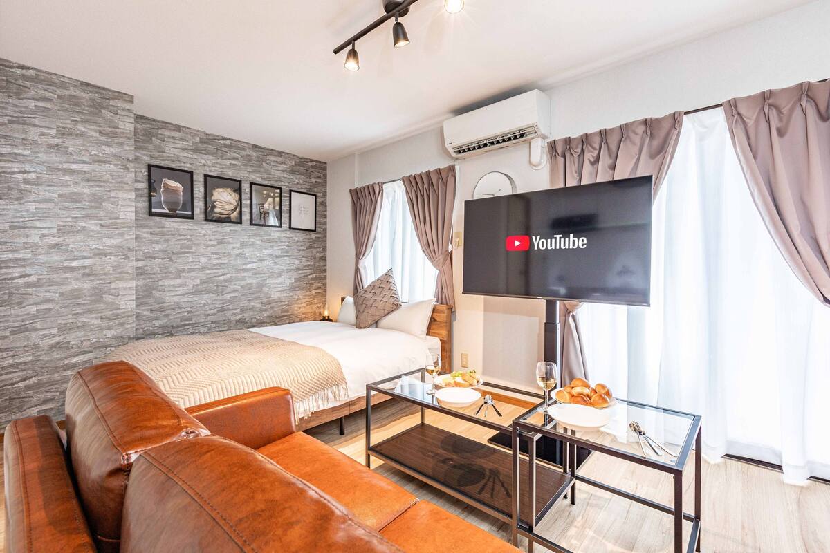 Quarto de hotel moderno com cama, mesa de centro de vidro, sofá de couro, televisão de tela plana e ar condicionado fixado na parede.