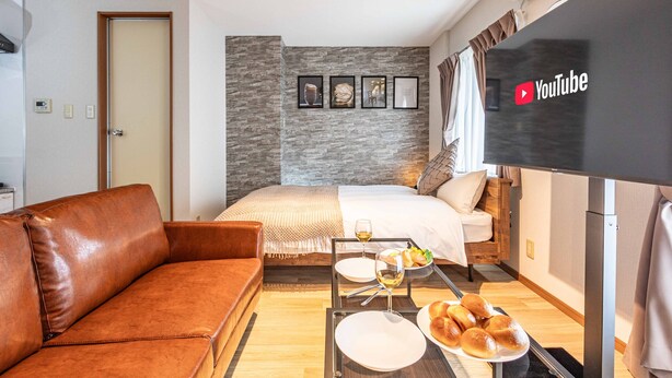 Une chambre d’hôtel moderne équipée d’un lit, d’un canapé en cuir marron, d’une table à manger avec des assiettes et des verres, et d’un téléviseur à écran plat affichant le logo de YouTube.