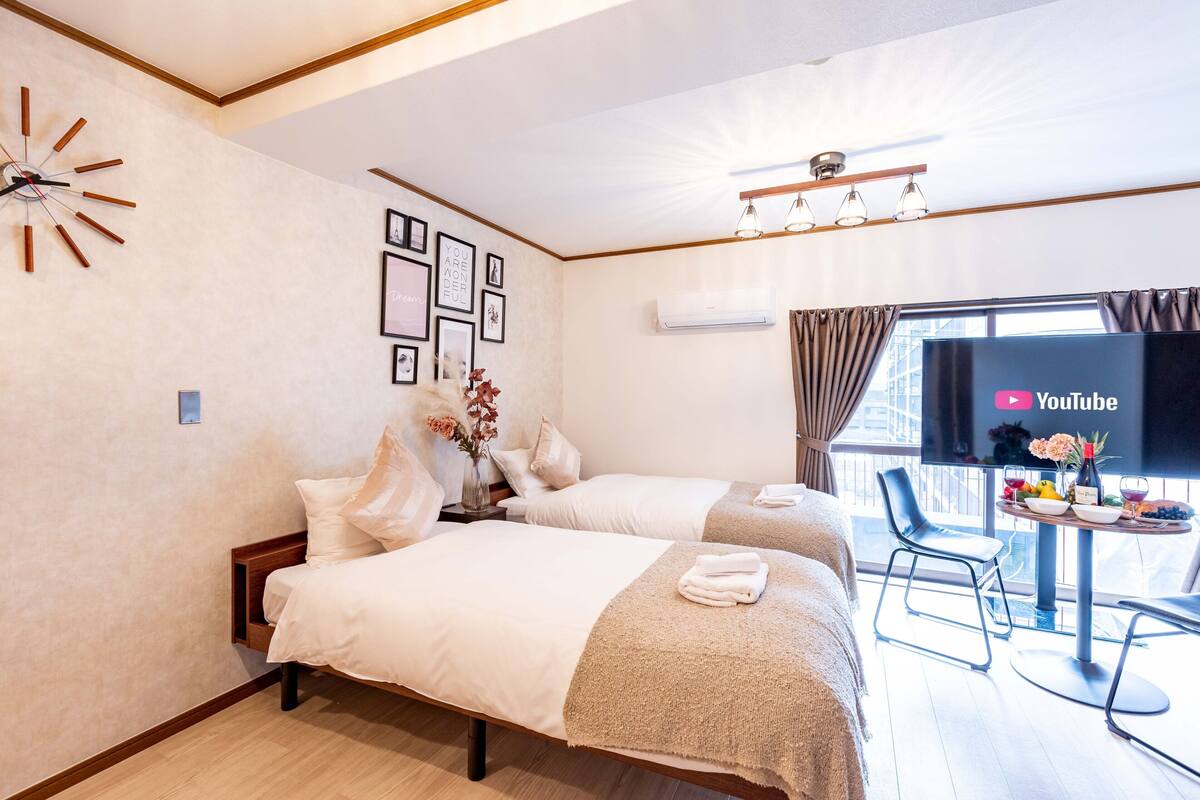 Quarto de hotel com duas camas, uma televisão exibindo YouTube, uma mesinha com um vaso e frutas, e uma janela com cortinas.