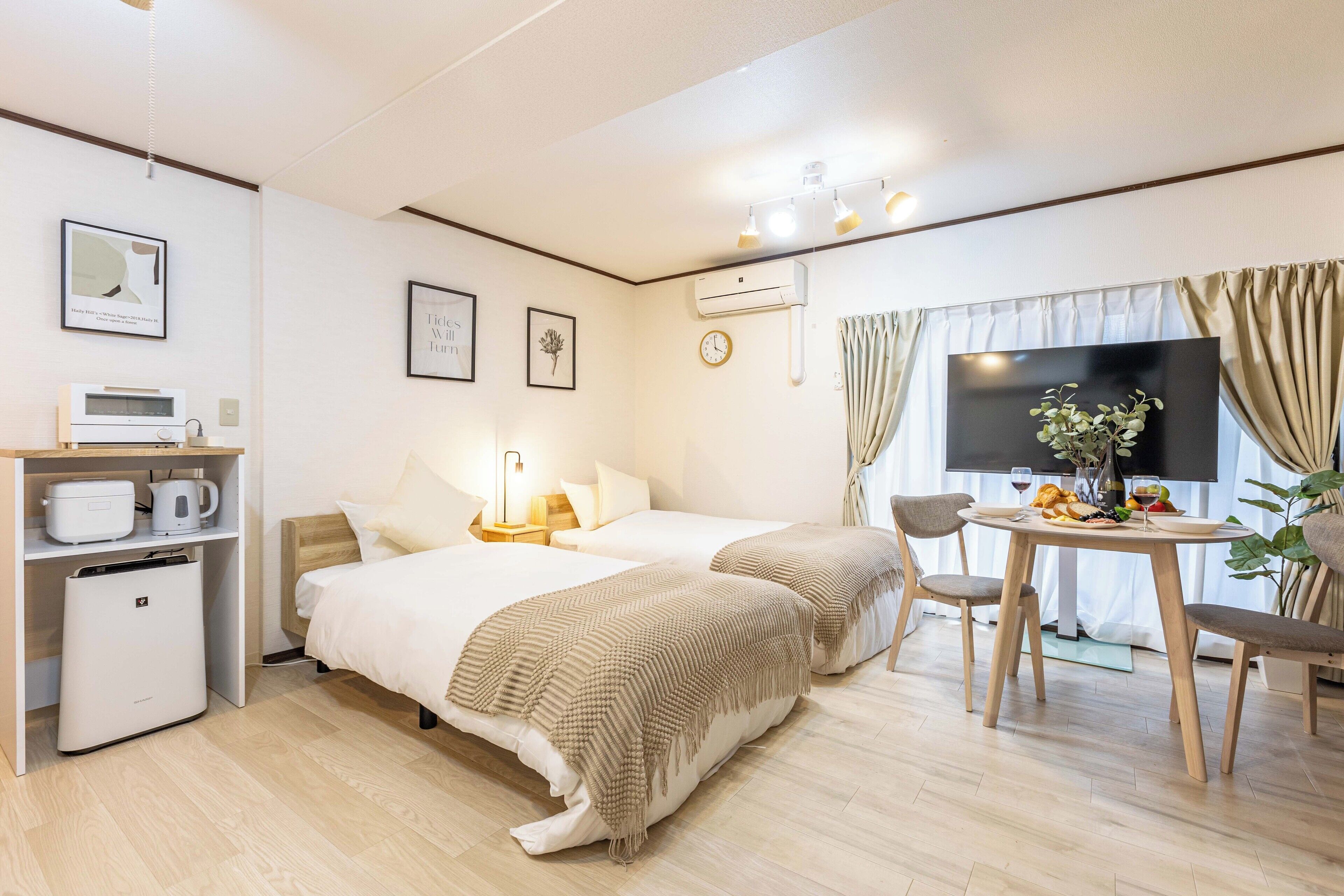 A hotel room with two beds, a small kitchenette, a dining table with chairs, a TV, and a plant.