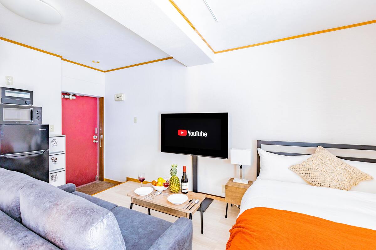 Quarto de hotel moderno com uma cama, televisão fixada na parede, área de refeições com mesa e cadeiras e uma kitchenette com frigorífico e micro-ondas.
