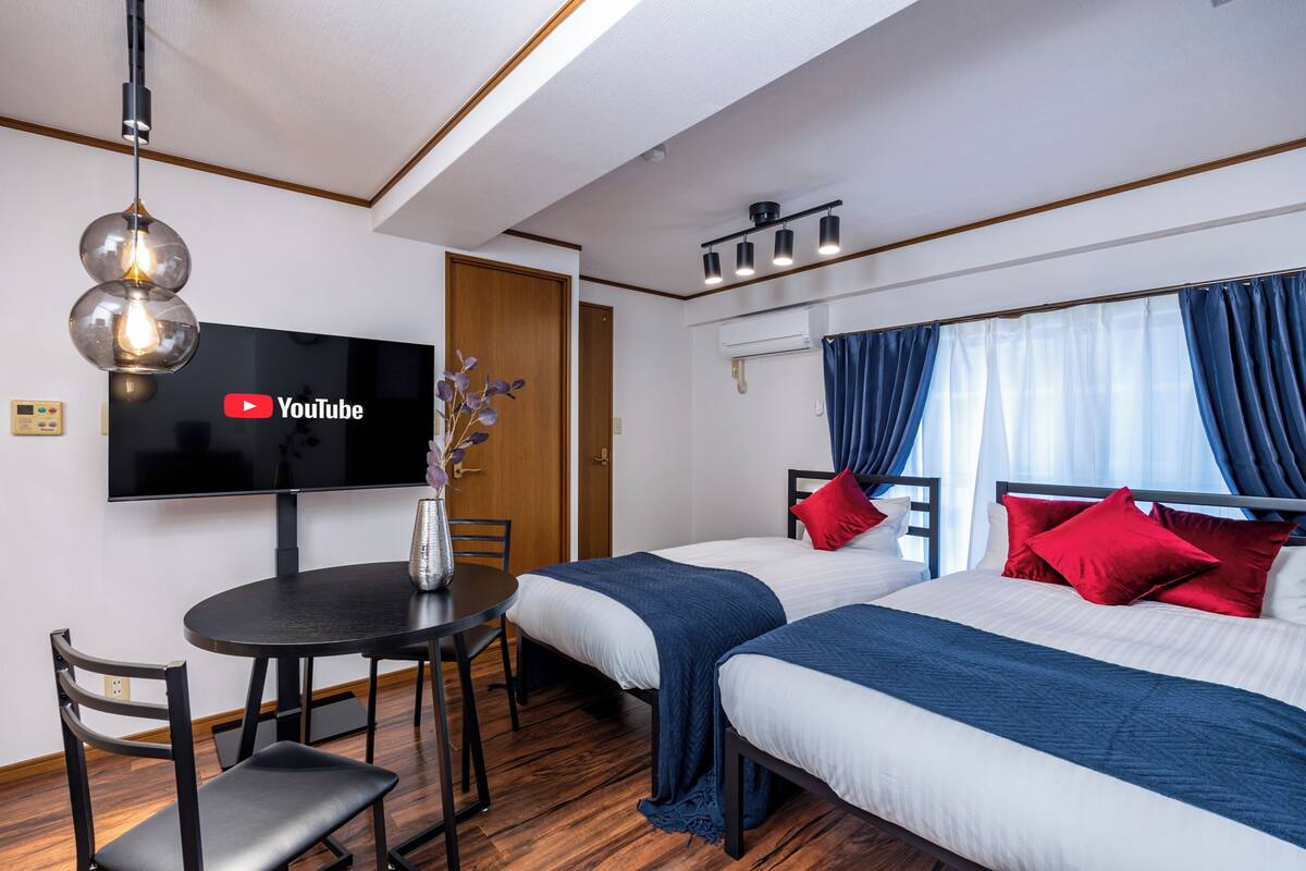 Quarto de hotel com duas camas, televisão, mesa de jantar com cadeiras e luminária no teto.