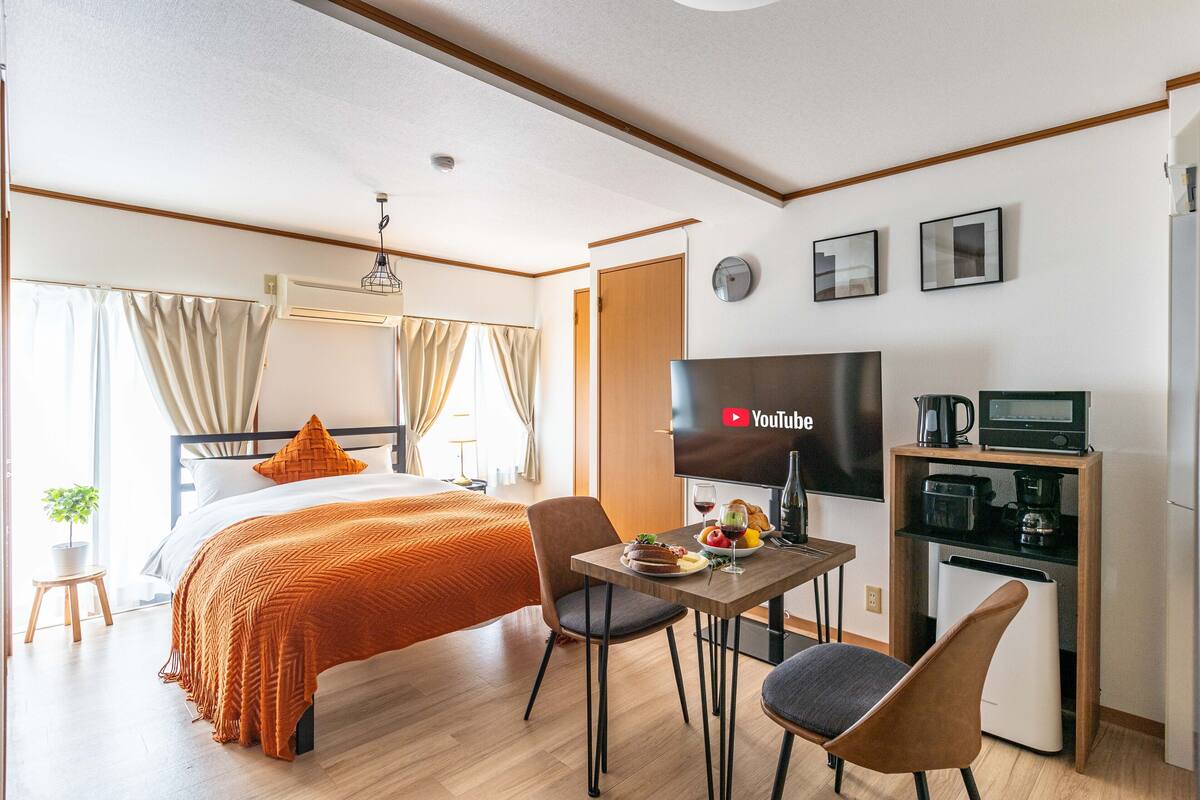 Quarto de hotel com cama, televisão, pequena mesa de jantar com um prato de comida, cadeira e cozinha compacta.