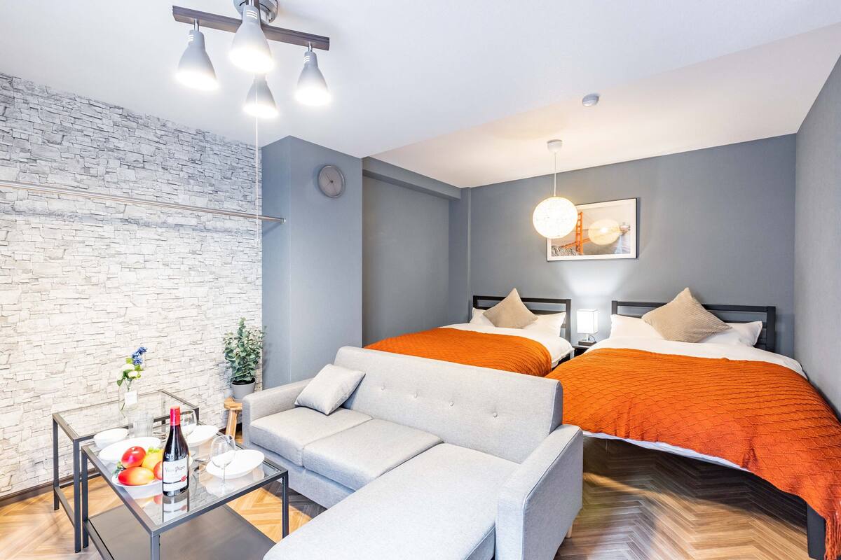 Quarto de hotel moderno com parede de pedra cinzenta, cama com roupa de cama laranja, sofá cinzento e mesa de centro de vidro com uma garrafa de vinho e fruta.