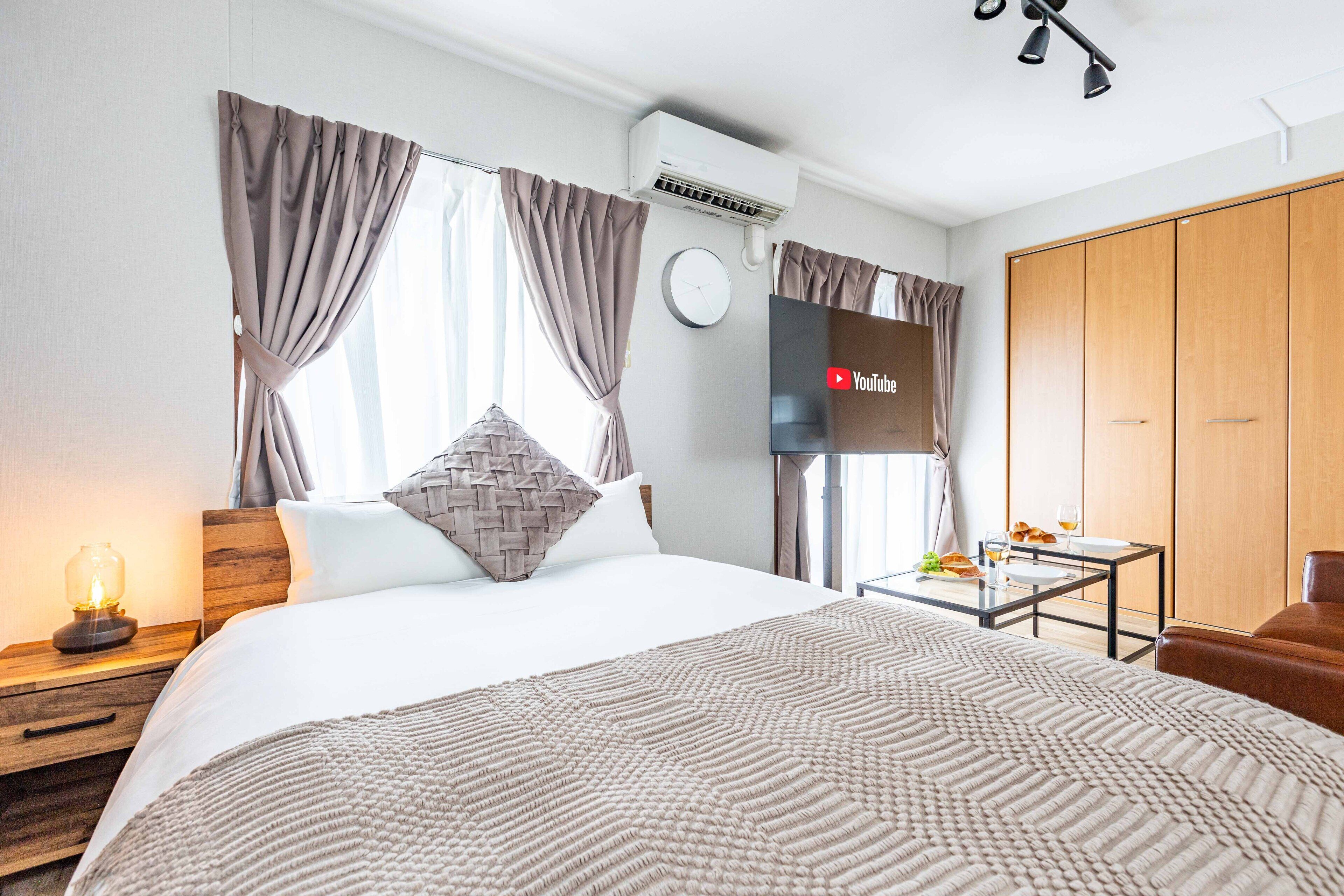 Una habitación de hotel moderna con una cama grande, una mesita de noche con lámpara, una pequeña mesa con artículos de desayuno y un armario.