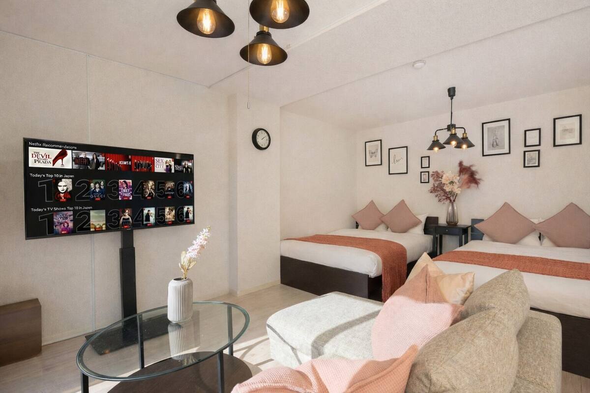 Quarto de hotel moderno com duas camas, uma mesa de centro de vidro, televisão fixada na parede exibindo cartazes de filmes e decoração minimalista.