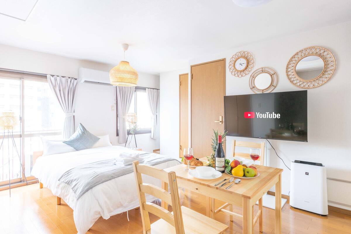 Quarto de hotel moderno com cama, mesa de jantar com frutas e vinho, televisão e um pequeno ar condicionado.