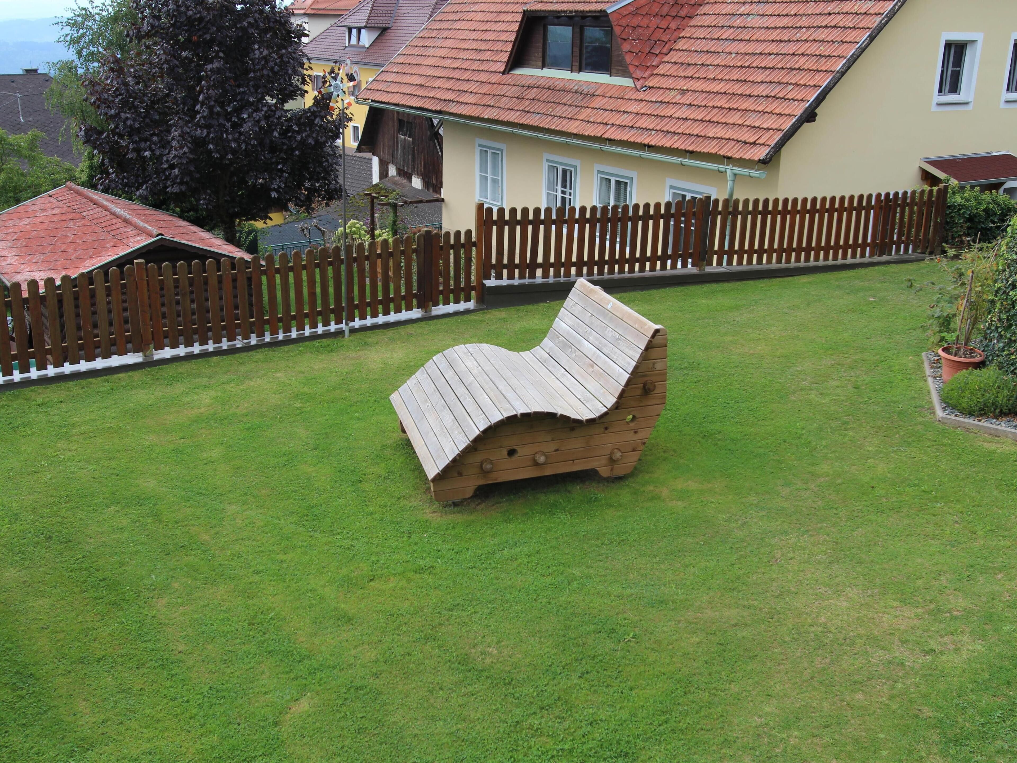 A wooden bench in a grassy yard with a wooden fence and a house with a red roof in the background.