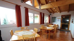 Un comedor con mesas y sillas de madera, ventanas amplias con cortinas y un suelo de baldosas.