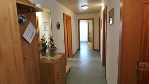 Un couloir avec des portes en bois, un banc, une petite table sur laquelle est posé un vase de fleurs et un panneau accroché au mur.
