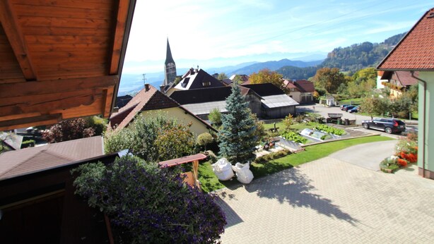 Une vue depuis une fenêtre donnant sur un village avec des maisons traditionnelles, une église et, au loin, une chaîne de montagnes.