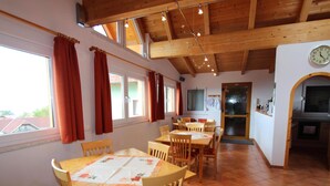Un coin repas avec des tables et des chaises en bois, une cuisine équipée d’un évier et d’une cuisinière, et une horloge au mur.