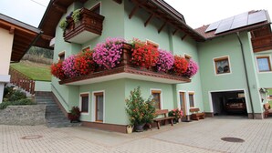 Edifici de dues plantes amb un balcó adornat amb flors de colors, una escalinata de pedra i un pati pavimentat.