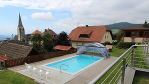 Una piscina amb una zona coberta, envoltada per una tanca de fusta, i una vista de cases i una església al fons.