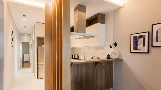 Une cuisine moderne avec des armoires en bois, une hotte de cuisinière en acier inoxydable et un four encastré. On y trouve un évier avec un robinet, une bouilloire et un vase décoratif contenant des feuilles. La cuisine est adjacente à un couloir menant à une salle de bain.