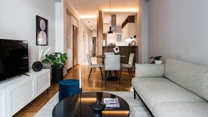 Un salon moderne doté d’un téléviseur à écran plat, d’un canapé, d’une table basse et d’un coin repas avec des chaises, et une cuisine en arrière-plan.