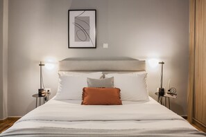 Ein ordentlich bezogenes Bett mit weißen Laken und einem braunen Kopfkissen, flankiert von zwei Nachttischen mit Lampen, einem gerahmten abstrakten Kunstwerk darüber und einer Holztür auf der rechten Seite.