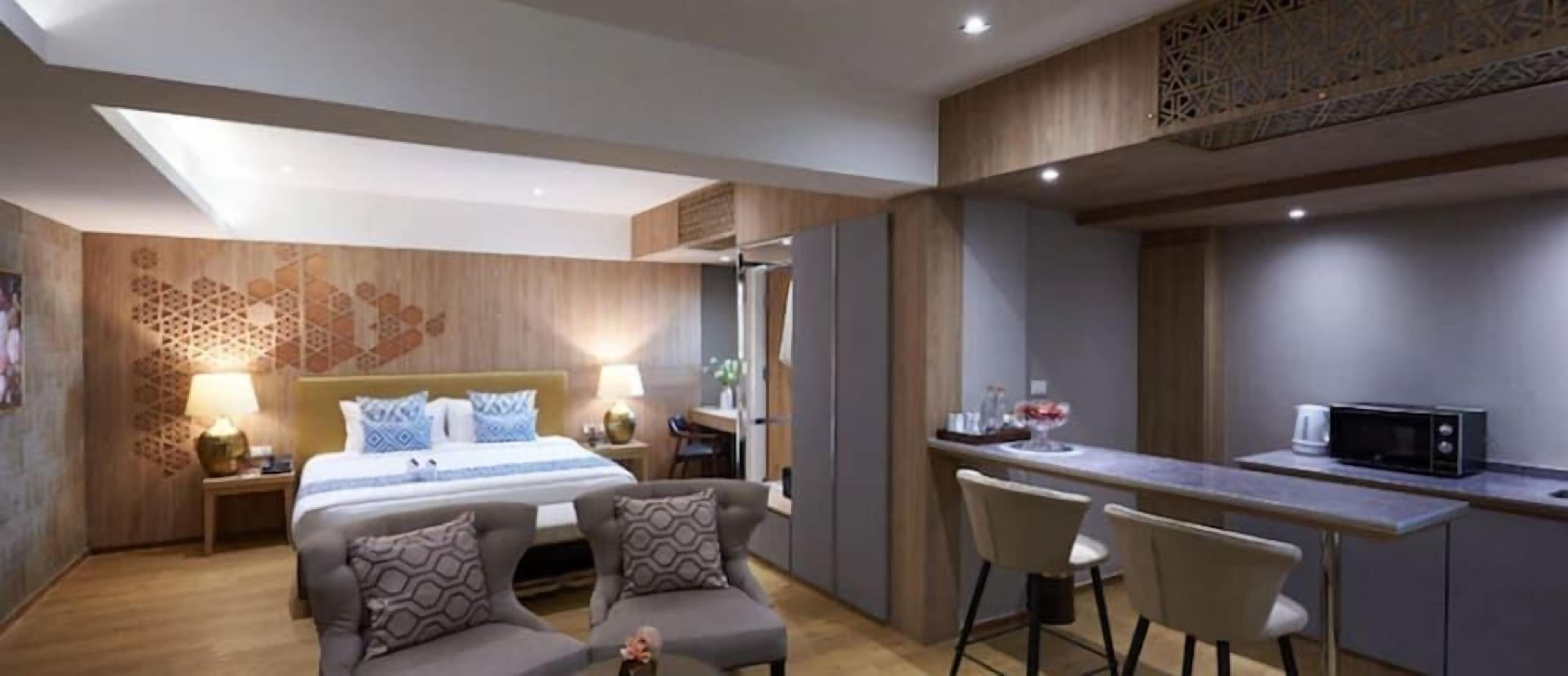 A modern hotel room with a bed, two armchairs, a small dining area with a microwave, and a decorative wall panel.