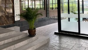 Entrada de um edifício moderno, com degraus de mármore, uma planta em vaso e grandes portas de vidro que dão para uma área externa.