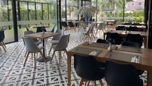 Uma área de refeições moderna com mesas, cadeiras, janelas amplas e vista para edifícios e vegetação.