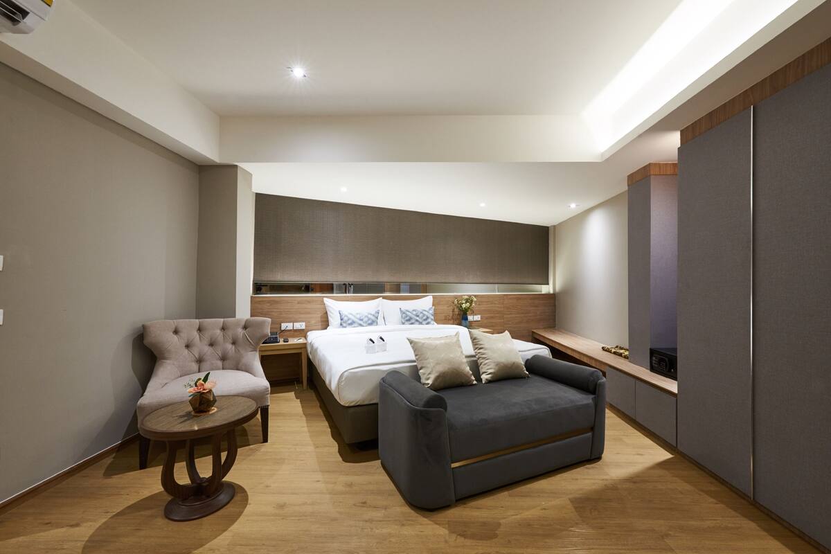 Quarto de hotel moderno com cama, sofá cinza, mesa de madeira redonda e uma janela grande.