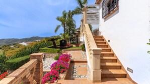 A staircase leading to a white building with a stone balustrade, surrounded by a landscaped garden with flowers and palm trees. - Villa Mirador de Frigiliana (Frigiliana)
