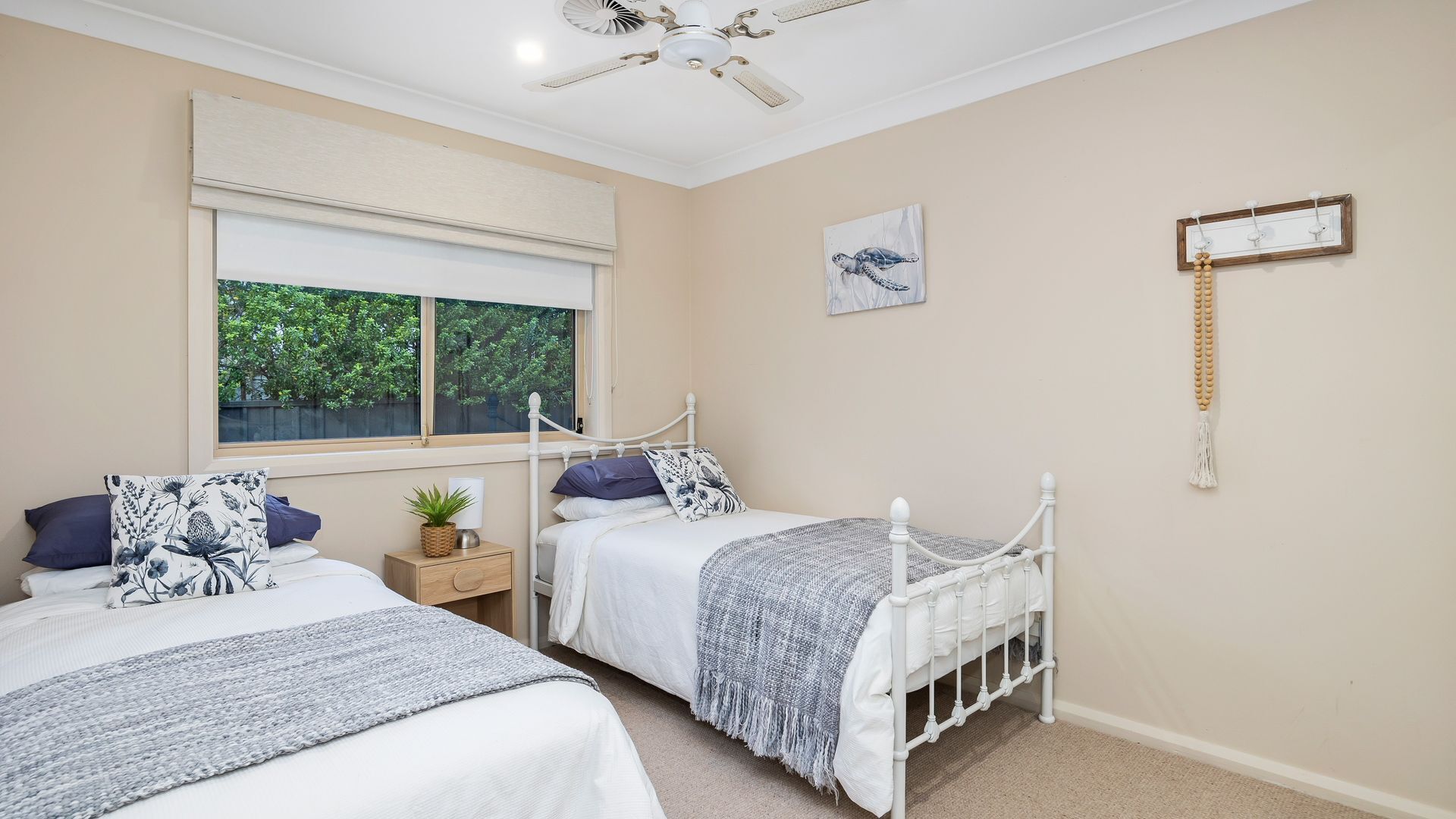 A bedroom with two beds, a ceiling fan, a window with a view of trees, a wall-mounted picture, and a bedside table with a plant.