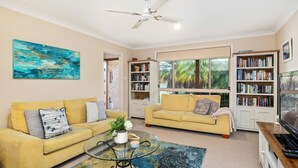 A living room with two yellow sofas, a glass coffee table, and a bookshelf filled with books. - Beachcomber - The Perfect Beach Getaway (Mollymook Beach)