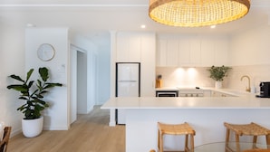 A modern kitchen with white cabinetry, a large island with stools, a hanging light fixture, and a potted plant. - Breakers 5 - Beautifully renovated apartment just a stones throw from the beach (Mollymook)