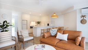 A modern living room with a brown leather sofa, a wicker chair, and a white coffee table with magazines. A kitchen with white cabinets and a hanging pendant light is visible in the background. - Breakers 5 - Beautifully renovated apartment just a stones throw from the beach (Mollymook)