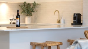 A modern kitchen with a white countertop, two wine glasses, a bottle of wine, a plant, a gold faucet, and a coffee machine. - Breakers 5 - Beautifully renovated apartment just a stones throw from the beach (Mollymook)