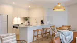 A modern kitchen with white cabinetry, a central island with stools, a coffee machine, and a hanging light fixture. - Breakers 5 - Beautifully renovated apartment just a stones throw from the beach (Mollymook)