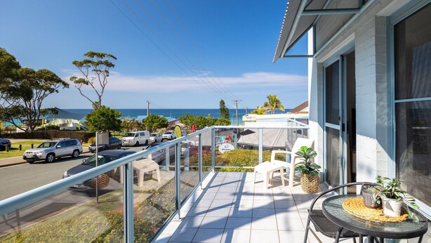 A balcony with a view of the ocean, a street with parked cars, and outdoor seating.