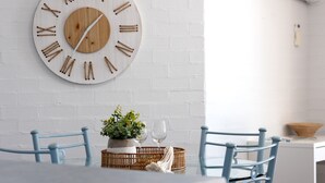 A dining area with a white brick wall, a wooden clock, two blue chairs, a table with a potted plant, and two wine glasses.