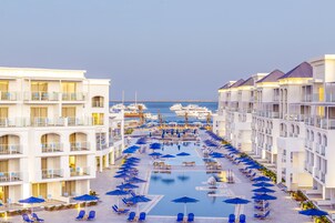 Un complejo turístico con una piscina central rodeada de sombrillas azules y sillones de descanso, varios pisos con balcones y vistas al mar.