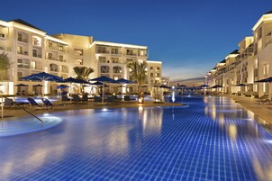 Zona de piscina de hotel con múltiples sillas de descanso y sombrillas, rodeada de edificios de varios pisos con balcones.